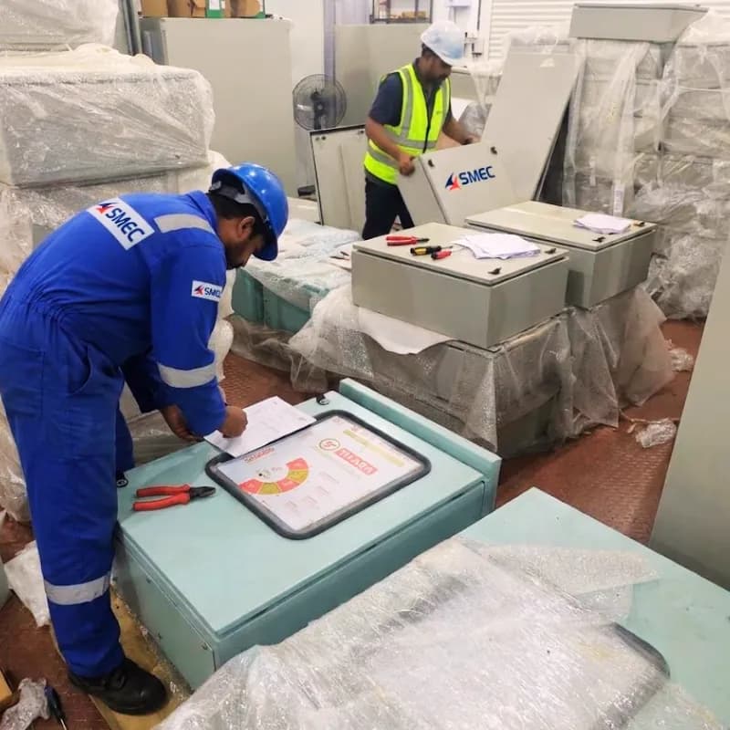 A technician in SMEC coveralls and a hard hat writing on a document beside wrapped control panels, with a colleague working behind him